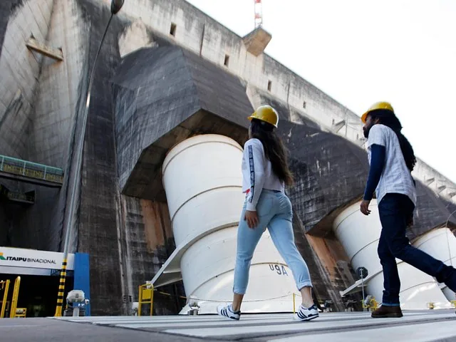 Two visitors in hard hats walking near massive penstocks at Itaipu Dam.