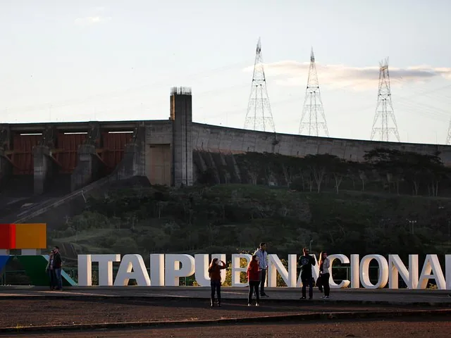 Tourists standing near large Itaipu Binacional sign with dam in background.