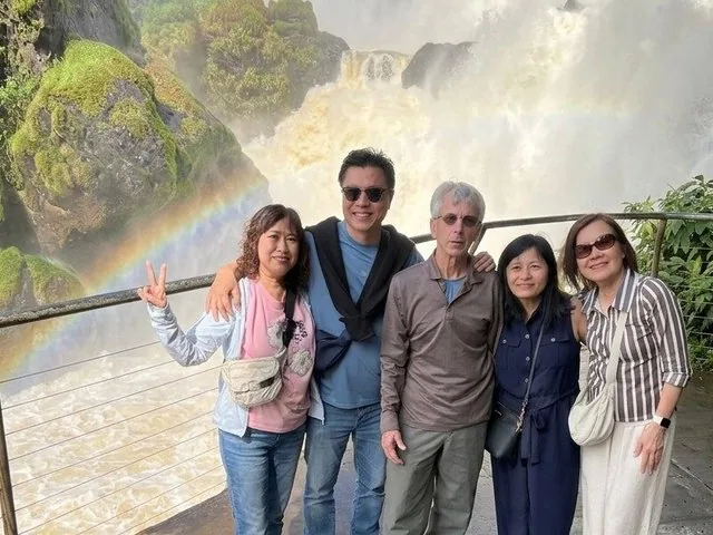 A group of five people posing in front of a waterfall with a rainbow.