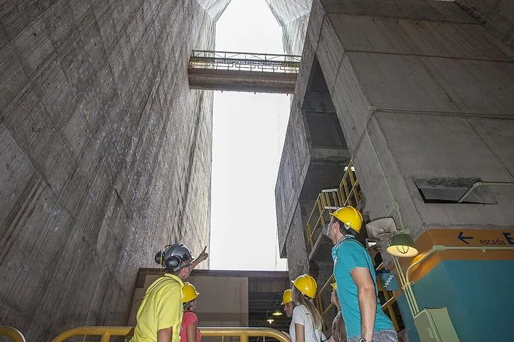 Tourists in hard hats looking up at the towering concrete walls of a dam.