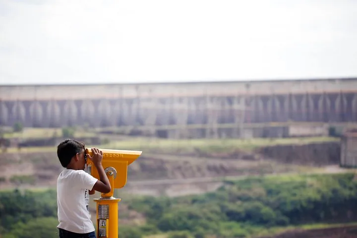 A child looking through a yellow telescope towards a large dam structure.