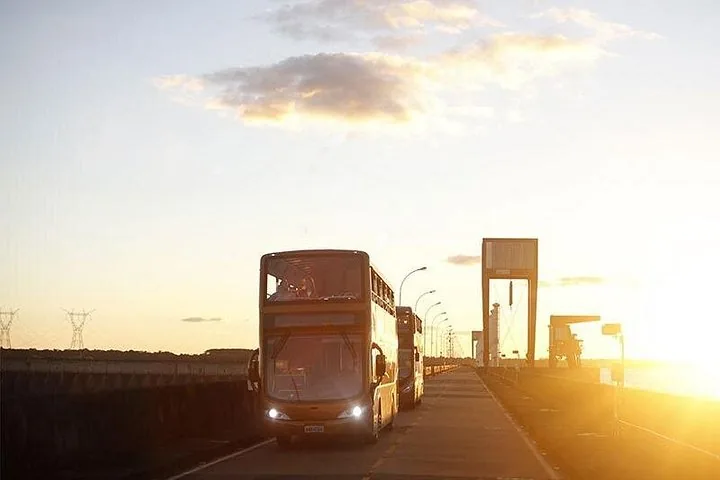 A double-decker bus driving along a road at sunset near Itaipu Dam.