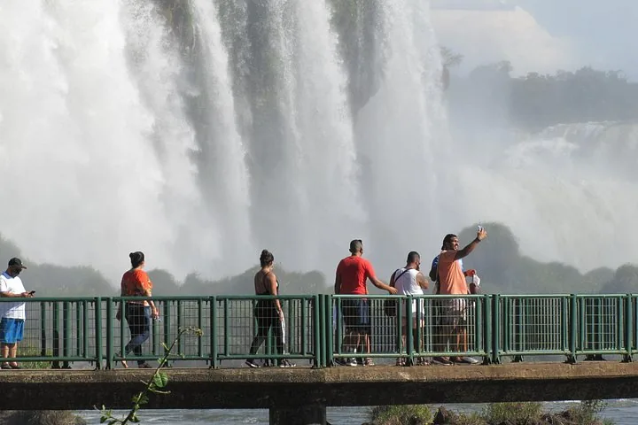 Tourists standing on a viewing bridge in front of massive, misty waterfalls.